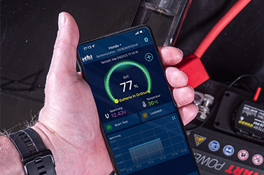 Das Bild zeigt ein Smartphone in der Hand vor einer Batterie. Darauf wird der SoC in der Battery-Guard App angezeigt.